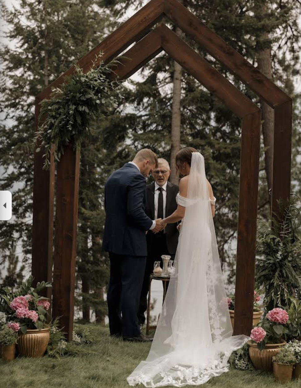 Bride and groom holding hands beneath a wooden arch during an outdoor wedding ceremony officiated by Celebrations by Stephen, surrounded by pine trees and floral arrangements.